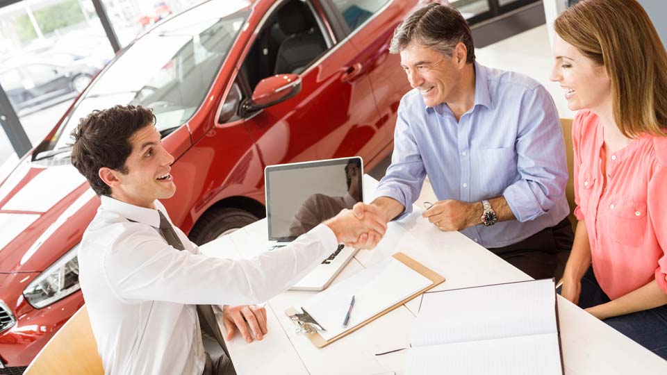 Ein Autoverkäufer schüttelt den Kunden die Hand in einem Autohaus in Butzbach, im Hintergrund ist ein rotes Auto zu sehen und auf dem Tisch liegen Unterlagen.